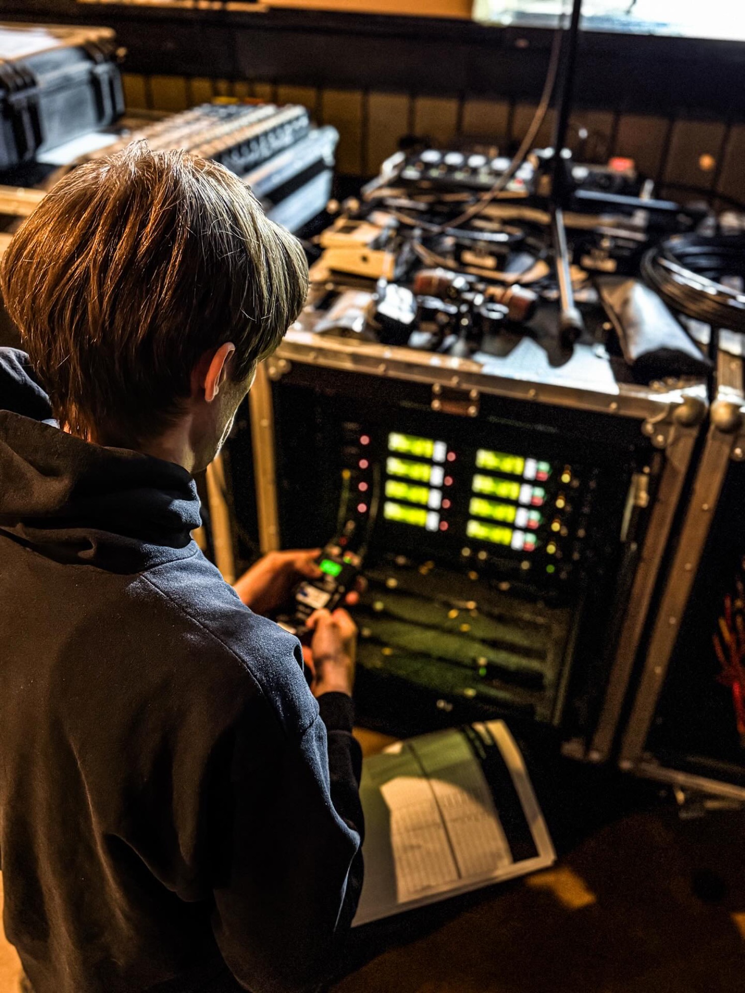 An engineer working a stage rack with green channel meters, comms in hand and a printed cue sheet, backstage.