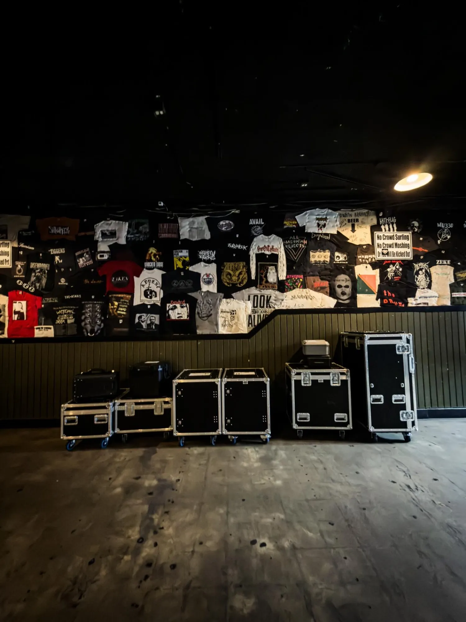 A row of road cases lined up on the floor of a music club, the back wall papered with band flyers, at load-in.