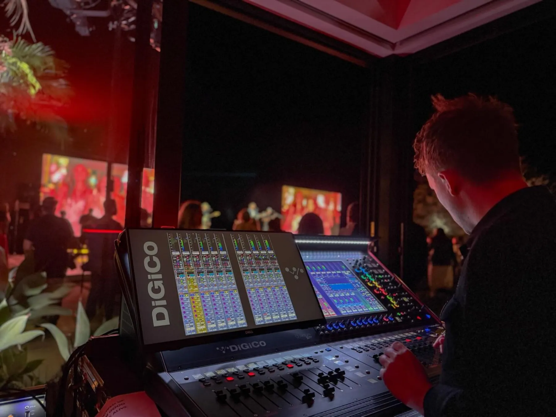 An over-the-shoulder view of an engineer mixing on a DiGiCo Quantum at a red-lit gala, blurred crowd beyond.