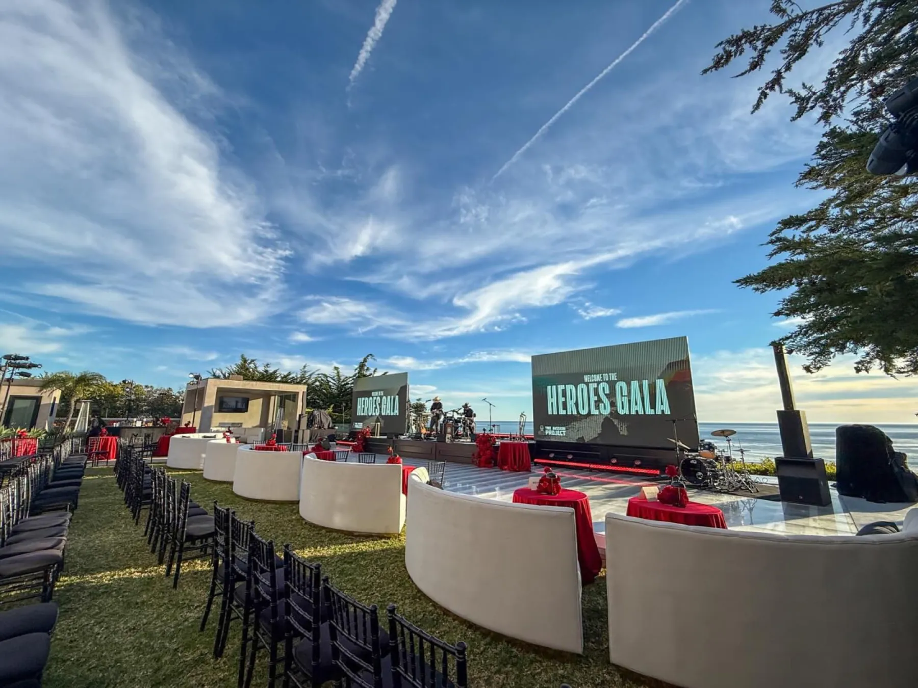 An outdoor oceanfront gala with a large LED stage screen, curved lounge seating, blue sky and the Pacific behind.