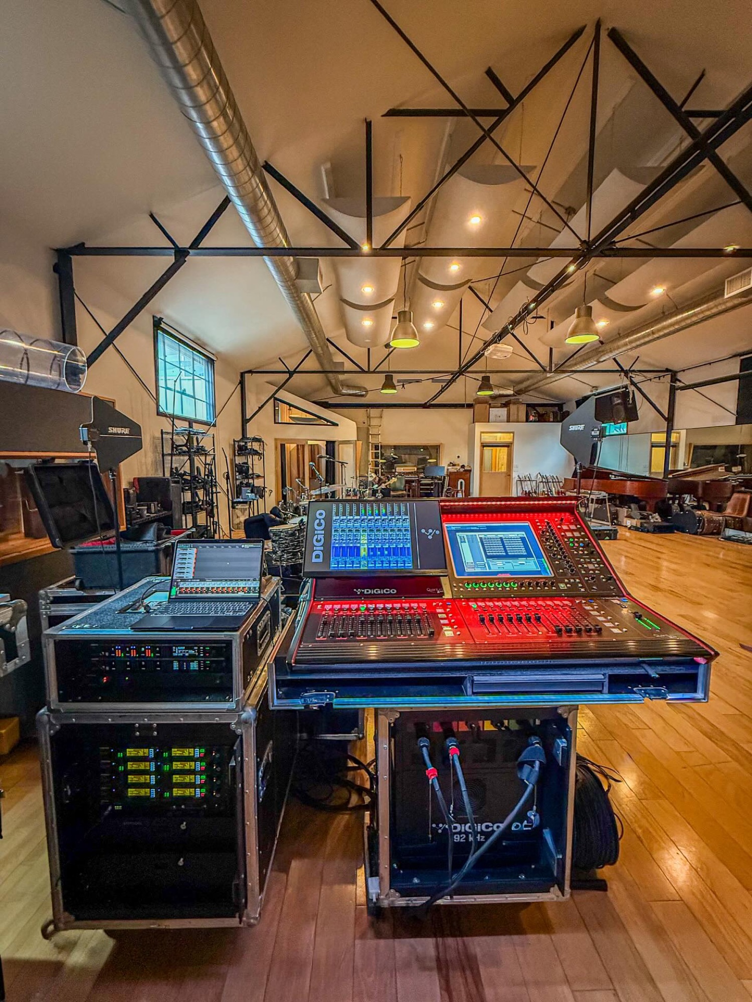 A front-of-house position with two DiGiCo consoles on cases in a high-ceilinged wood-floor hall, a drum kit on the stage behind.