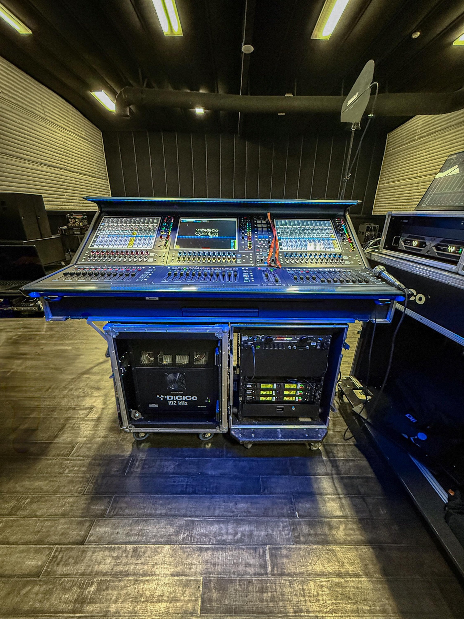 A blue-lit DiGiCo SD console on cases with stage racks below, in a dark venue with hanging choir microphones.