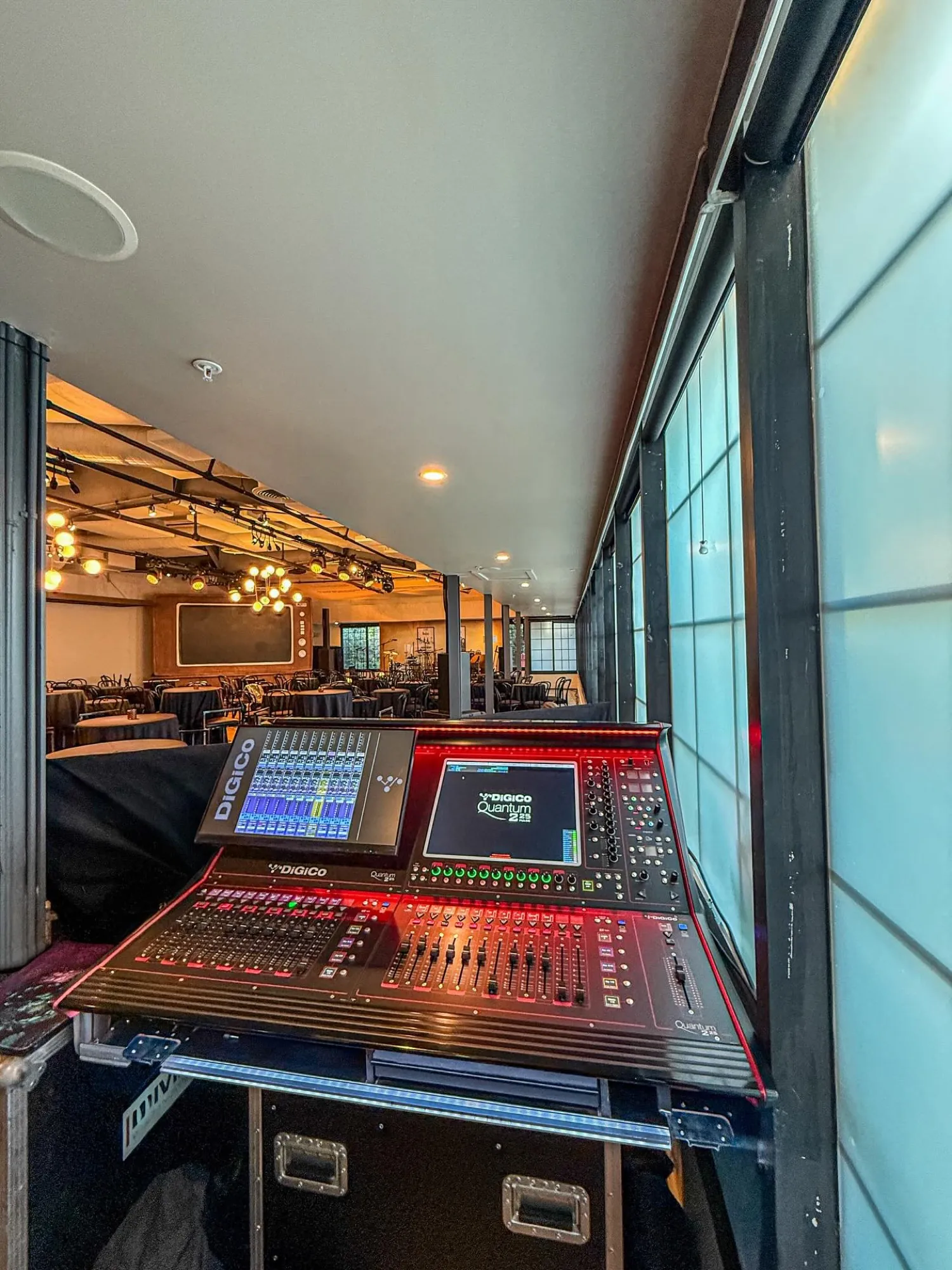 A red-lit DiGiCo Quantum at a raised front-of-house booth beside frosted-glass windows overlooking an event hall.