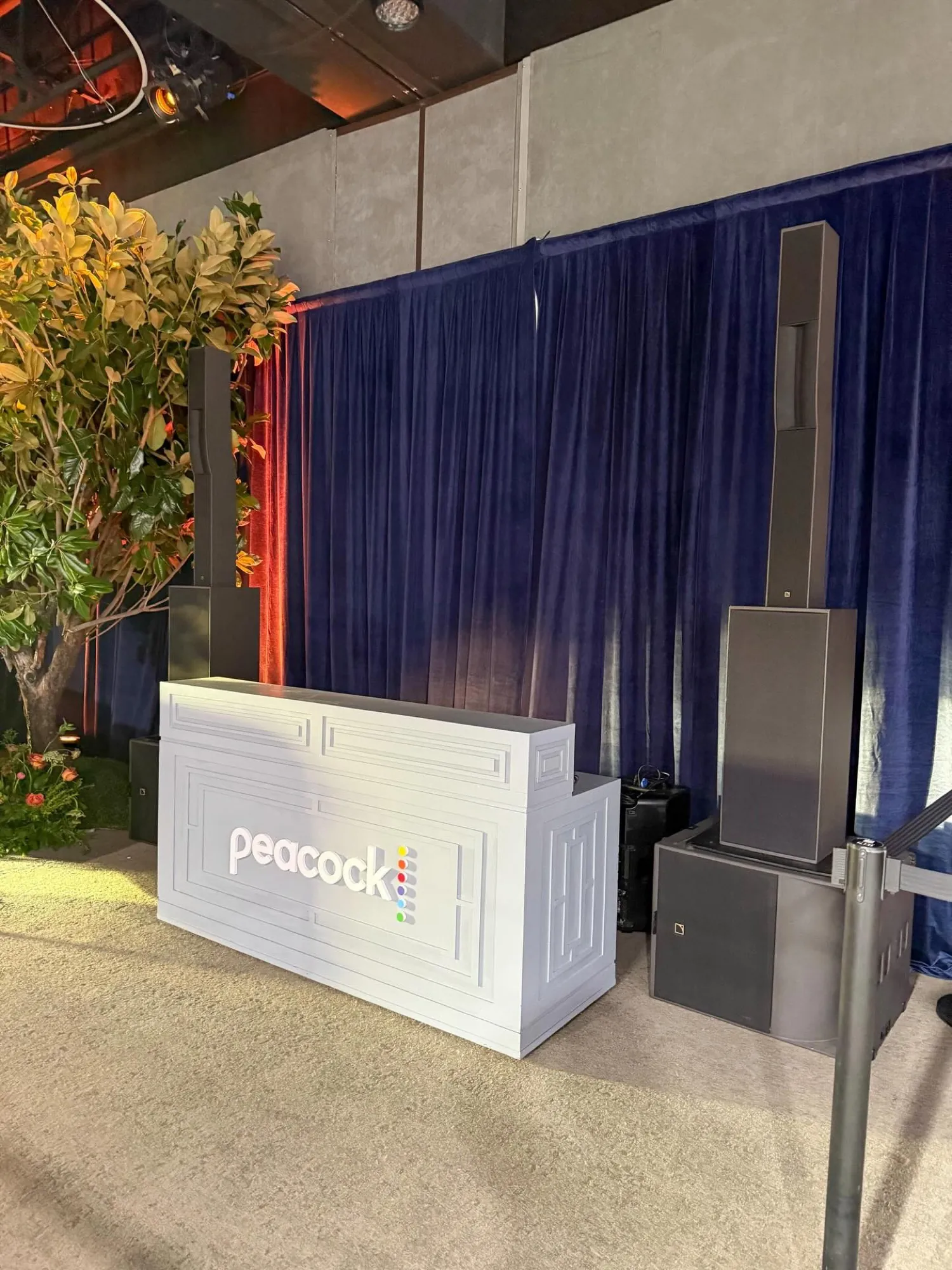 A white-branded control booth flanked by line-array speakers and a subwoofer against blue drape at an outdoor brand activation.