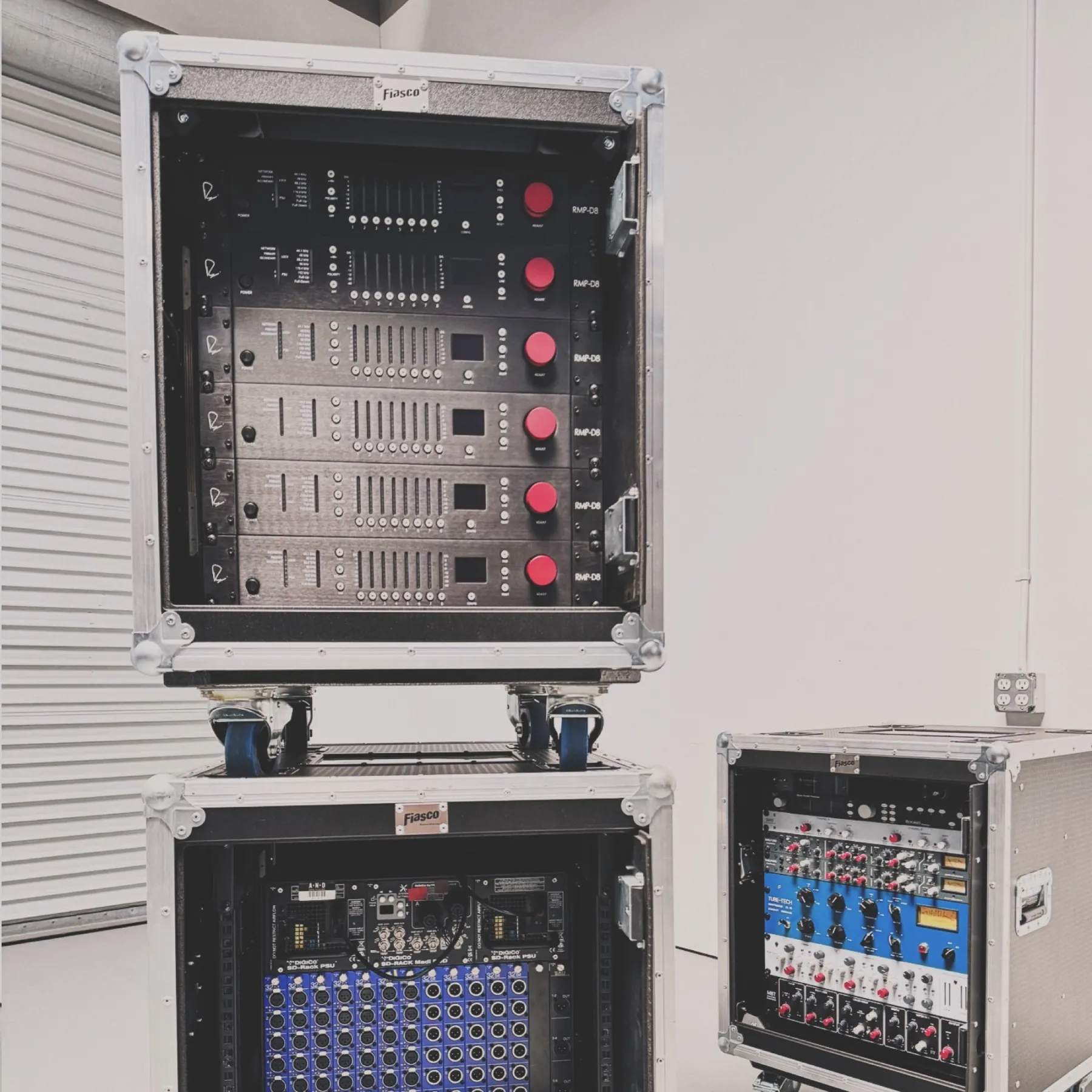 An open rack of power amplifiers stacked on a patch rack with a second rack alongside, warehouse roll-up door in frame.